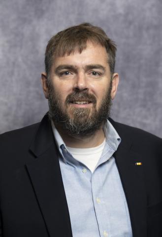 Headshot of Josh Cannon. He has a beard and is wearing a blue collared shirt and black jacket. 