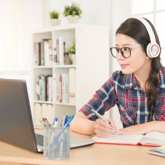 person with headphones on, writing in a notebook while looking at laptop screen